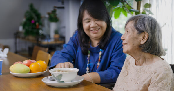 Caregiver sitting at table, feeding elder adult